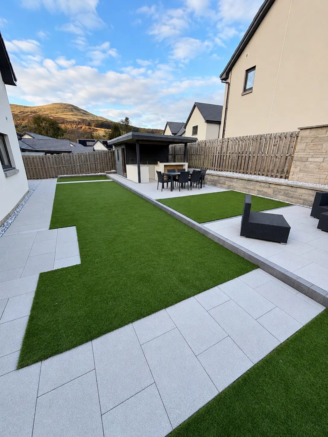 New build garden with artificial grass lawn, porcelain paved patio and outdoor kitchen with dining area under blue skies