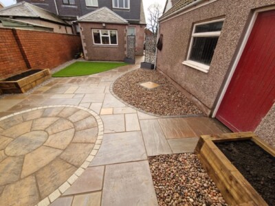 "Transform your outdoor space with a stunning patio design. This unique installation features a beautiful Indian sandstone patio with a Tobermore historic circle, accompanied by artificial grass, timber sleepers, and anthracite wall cladding. Create a captivating and versatile outdoor oasis for relaxation and entertaining." A stunning patio installation showcasing a unique combination of materials. A natural stone Indian sandstone patio serves as the foundation, exuding elegance and timeless charm. A Tobermore historic circle adds a touch of intricate design, creating a focal point within the space. Artificial grass is seamlessly integrated, providing a lush and low-maintenance area for relaxation and play. Timber sleepers border the patio, adding a rustic and organic element, while anthracite wall cladding brings a modern and sleek touch to the overall aesthetic. This patio installation harmoniously blends natural stone, artificial grass, timber sleepers, and anthracite wall cladding, resulting in a visually captivating and versatile outdoor space. Garden Landscaping Edinburgh, Garden Landscaping Alloa, Garden Rooms Edinburgh, Garden Rooms Alloa