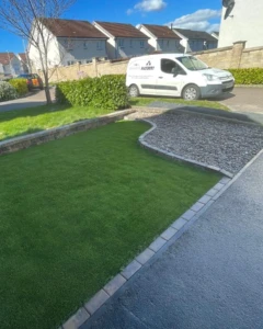 "Transformed and maintenance-free! Check out this stunning garden makeover in Tullibody, Alloa. We installed artificial grass and Scottish pebble stones, creating a sleek and modern look. The boundary is beautifully defined with a Tobermore Bracken wall and Bracken kerbstone. Say goodbye to garden chores and hello to contemporary elegance!" "Front garden design in Alloa featuring low-maintenance artificial grass and Scottish pebble stones. The boundary is defined by a Tobermore Bracken wall and Bracken kerbstone."