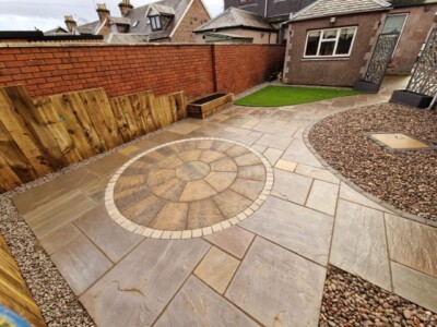 "Transform your outdoor space with a stunning patio design. This unique installation features a beautiful Indian sandstone patio with a Tobermore historic circle, accompanied by artificial grass, timber sleepers, and anthracite wall cladding. Create a captivating and versatile outdoor oasis for relaxation and entertaining." A stunning patio installation showcasing a unique combination of materials. A natural stone Indian sandstone patio serves as the foundation, exuding elegance and timeless charm. A Tobermore historic circle adds a touch of intricate design, creating a focal point within the space. Artificial grass is seamlessly integrated, providing a lush and low-maintenance area for relaxation and play. Timber sleepers border the patio, adding a rustic and organic element, while anthracite wall cladding brings a modern and sleek touch to the overall aesthetic. This patio installation harmoniously blends natural stone, artificial grass, timber sleepers, and anthracite wall cladding, resulting in a visually captivating and versatile outdoor space. Garden Landscaping Edinburgh, Garden Landscaping Alloa