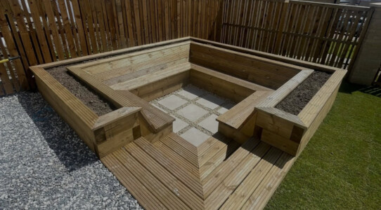 "Immerse yourself in the beauty of nature with our mesmerizing sunken garden installation, featuring exquisite porcelain tile and cozy timber seating. Experience tranquility in the heart of Dunfermline." A sunken area with porcelain tile surrounded by timber seating area and plant beds, installed in Dunfermline. Turf also installed.